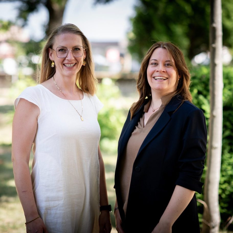 Zwei Frauen lächeln in einem grünen Garten. Beide tragen elegante Kleidung.