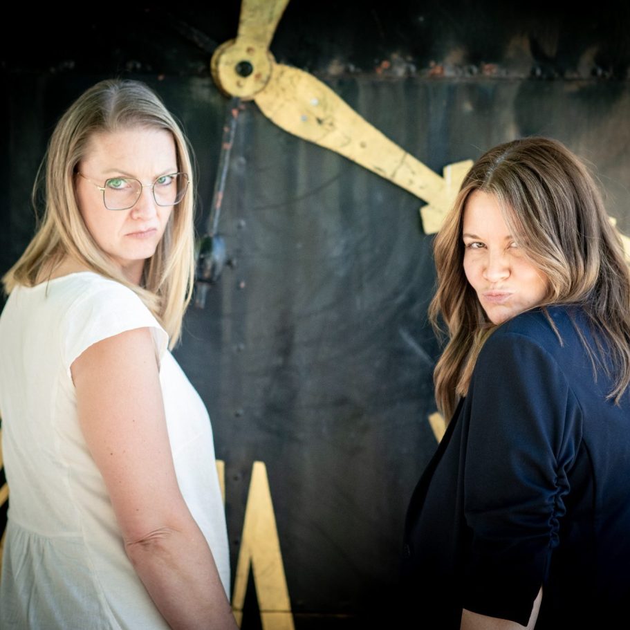 Zwei Frauen posieren vor einer großen Uhr mit einem geheimnisvollen Blick.