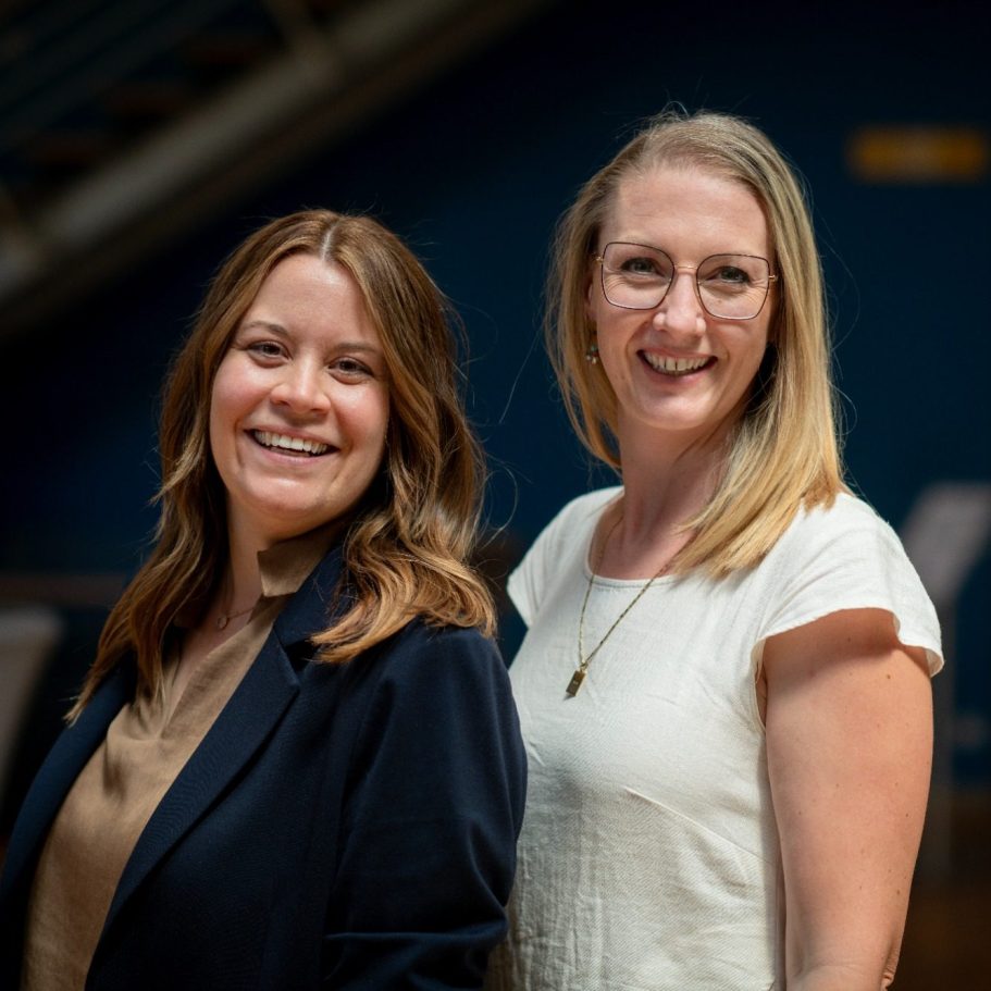 Zwei lächelnde Frauen stehen nebeneinander in einem modernen Innenraum.