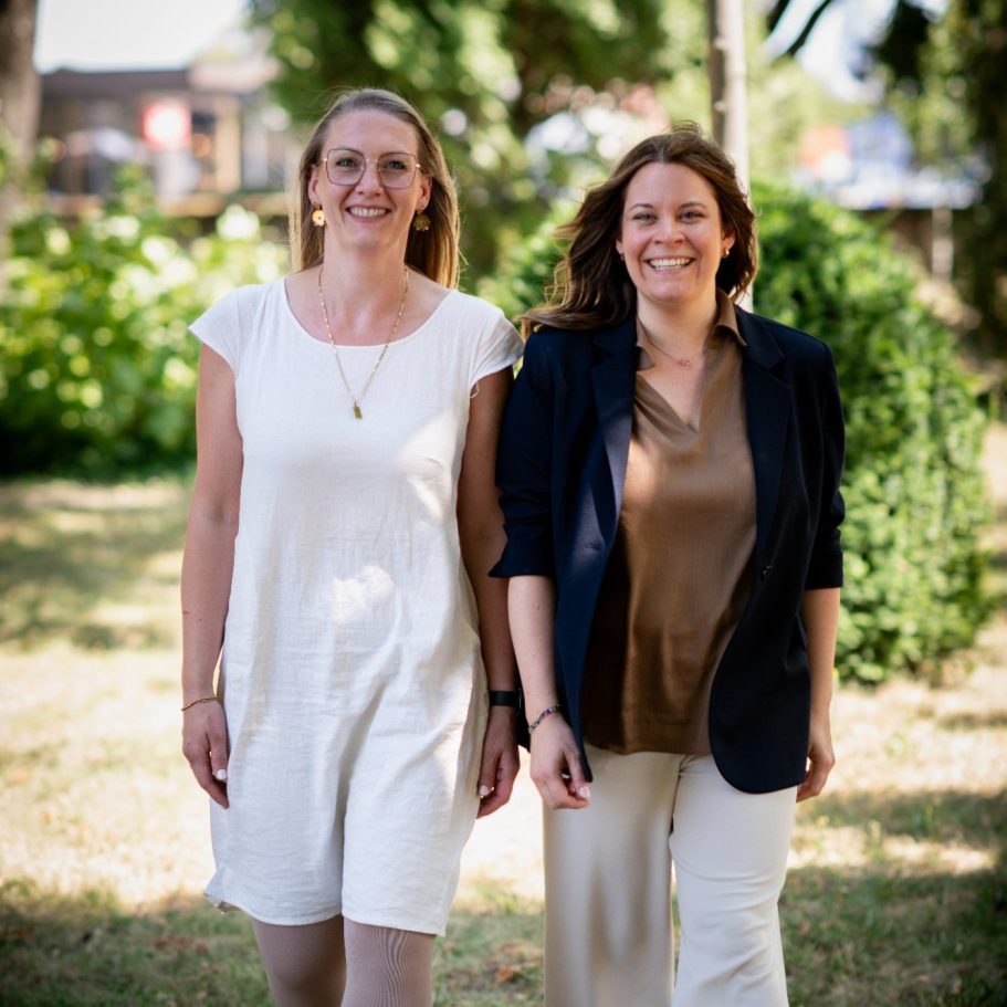 Zwei Frauen in entspannter Kleidung gehen lächelnd in einem grünen Garten.