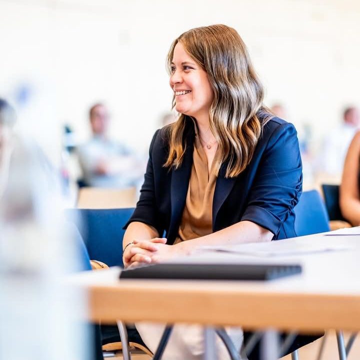 Frau mit lässiger Frisur und Blazer, lächelnd in einer Konferenzumgebung.