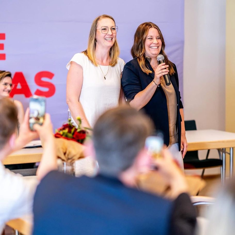 Zwei Frauen sprechen am Mikrofon, während das Publikum mit Handys filmt.