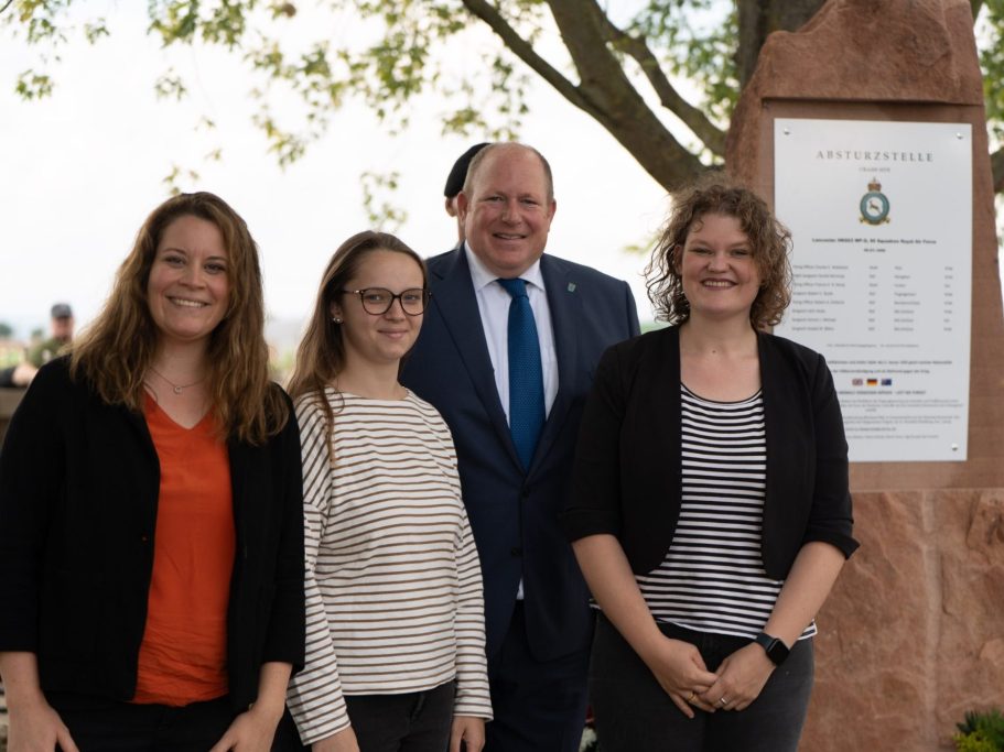 Vier Personen stehen lächelnd vor einer Gedenktafel im Freien.