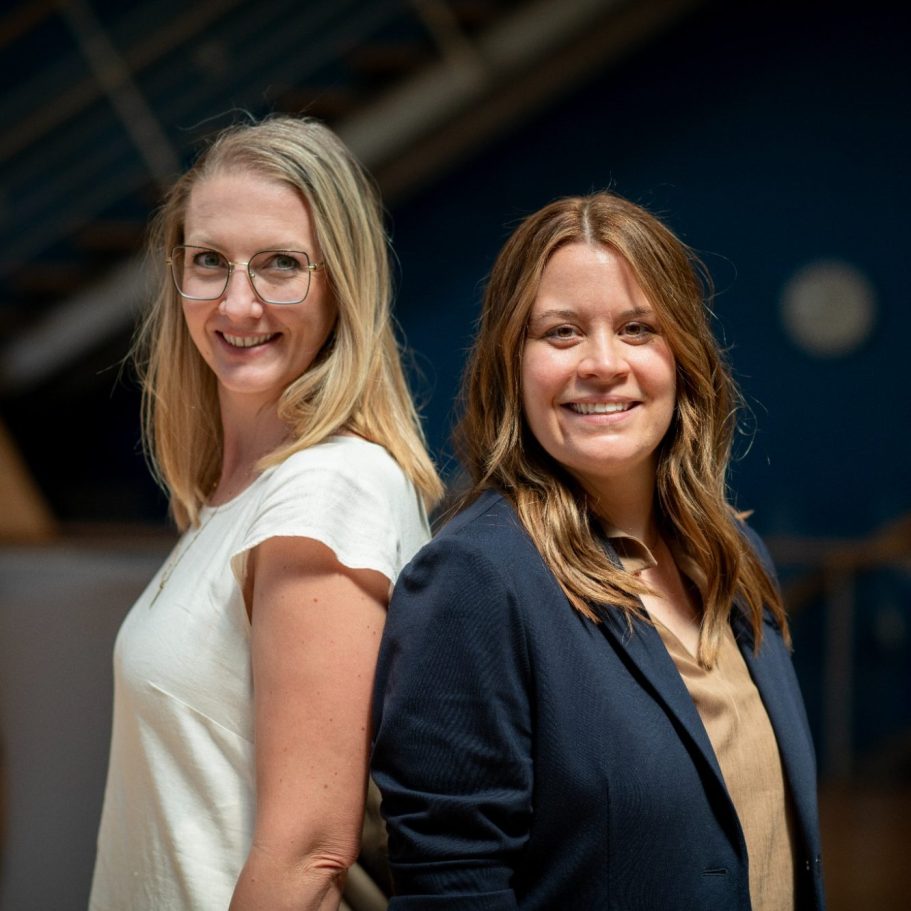 Zwei Frauen stehen Rücken an Rücken und lächeln in die Kamera.