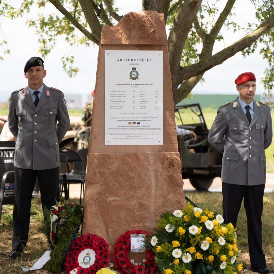 Zwei Soldaten neben einem Ehrenmal, umgeben von Blumen und einer Militärfahrzeug im Hintergrund.
