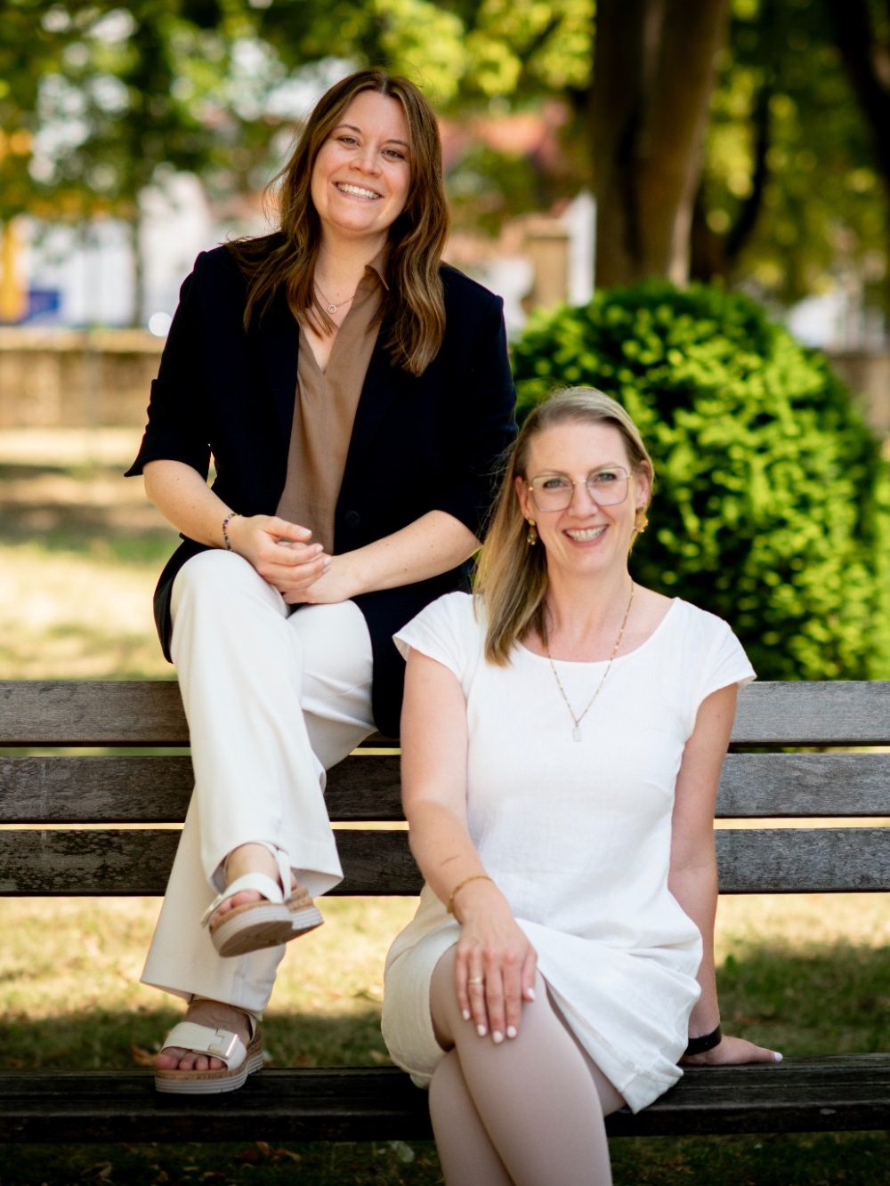 Zwei Frauen sitzen auf einer Bank in einem Park, umgeben von grünen Büschen.